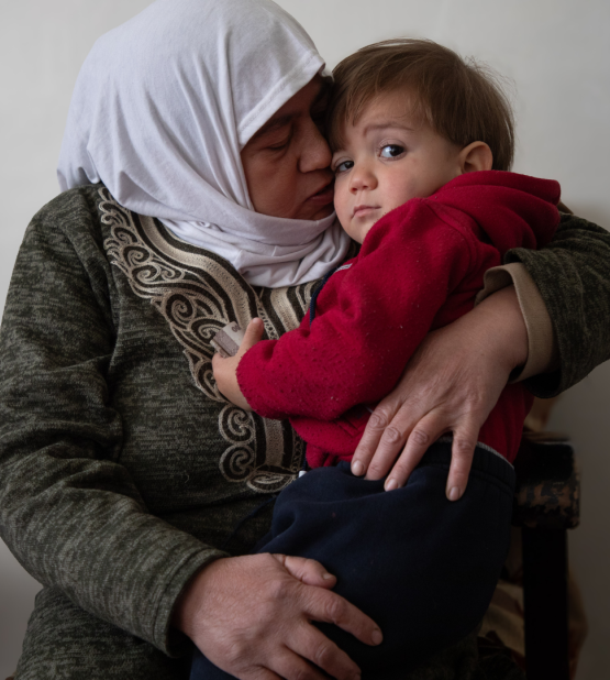 Fatmeh cassrying her grandson while sitting on a sofa inside her house in Hanano neighborhoud.