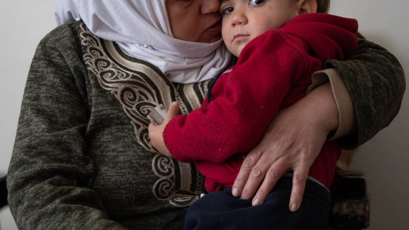Fatmeh cassrying her grandson while sitting on a sofa inside her house in Hanano neighborhoud.